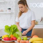Woman in white t-shirt preparing fresh vegetables in a bright kitchen with a laptop.